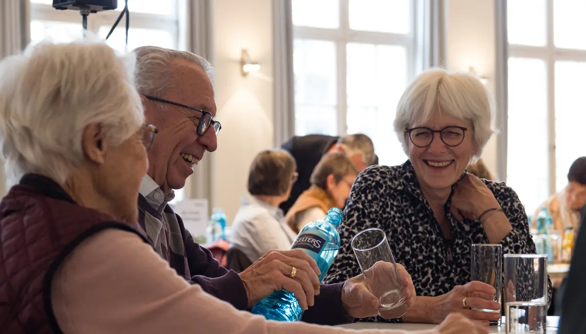 Zwei Frauen, ein Mann mit weißen Haaren sitzen an einem Tisch und lachen. | © Mick Murphy