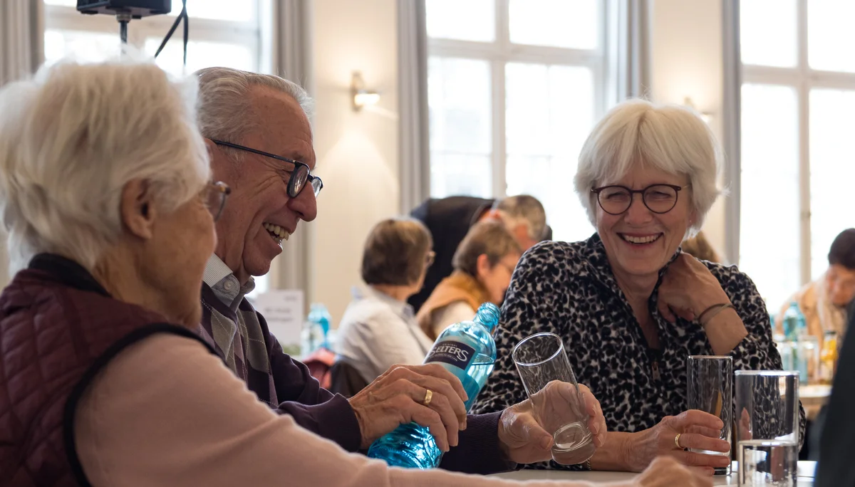 Zwei Frauen, ein Mann mit weißen Haaren sitzen an einem Tisch und lachen. | © Mick Murphy