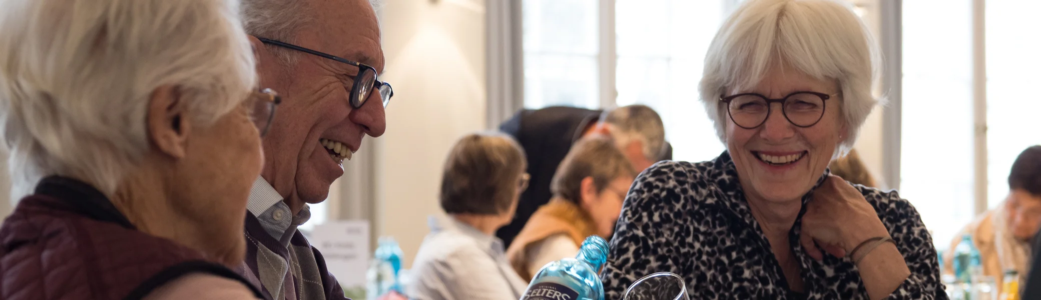 Zwei Frauen, ein Mann mit weißen Haaren sitzen an einem Tisch und lachen. | © Mick Murphy