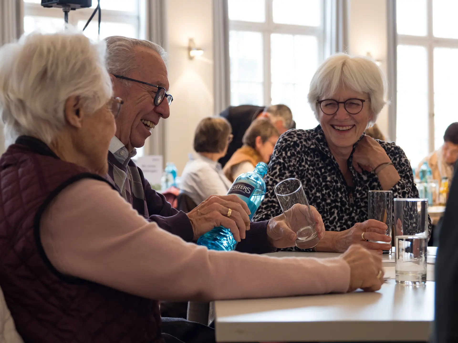 Zwei Frauen, ein Mann mit weißen Haaren sitzen an einem Tisch und lachen. | © Mick Murphy