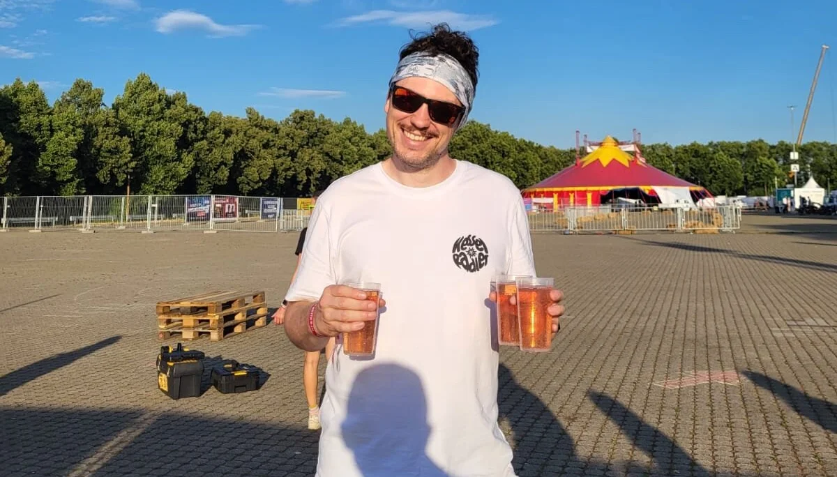 Mann mit weißem T-Shirt hält orangene Getränkebecher in der Hand | © Gabriel Molitor