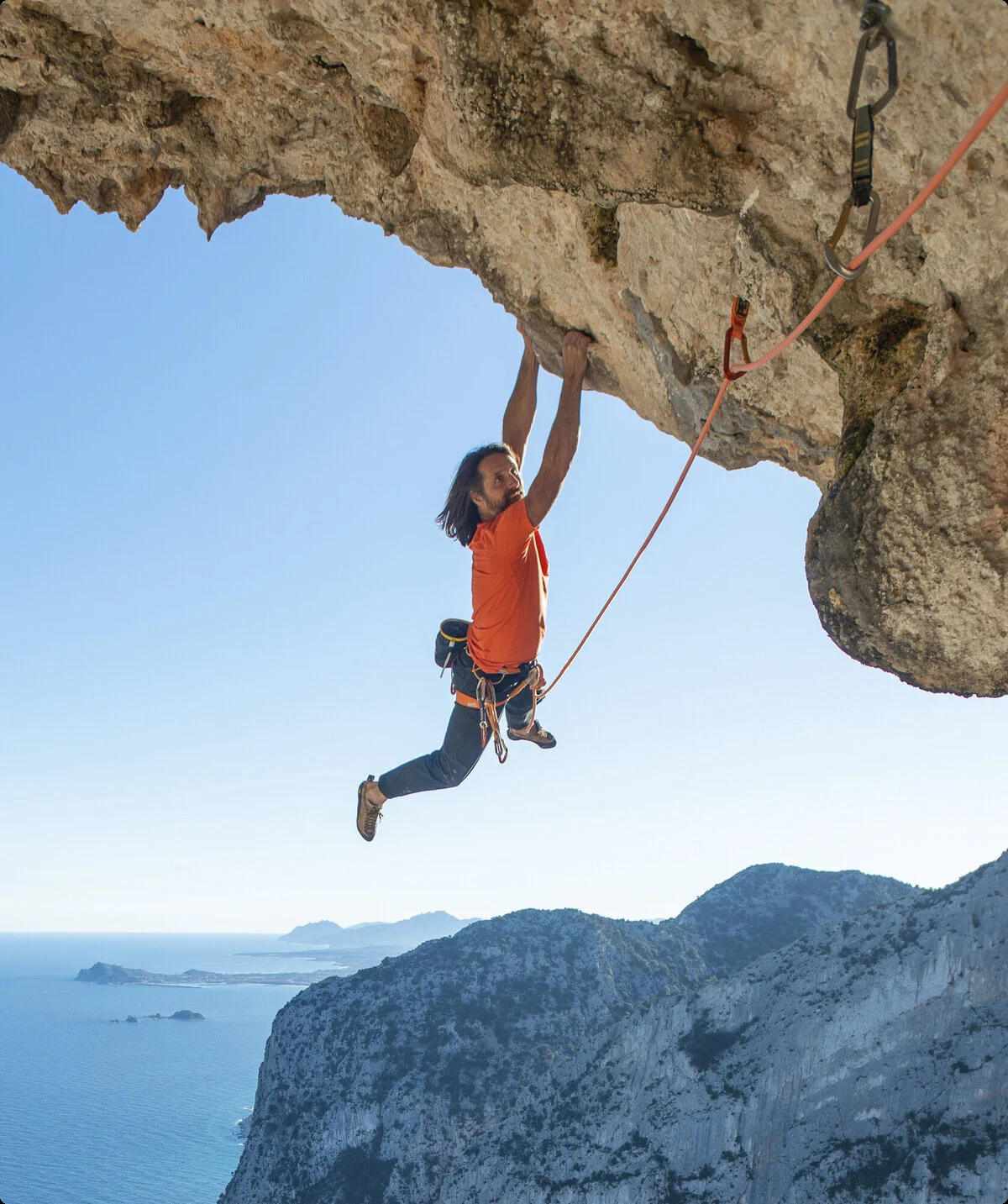 Mann hängt nur an den Armen im Überhang, im Hintergrund sieht man das Meer | © Alexander Huber