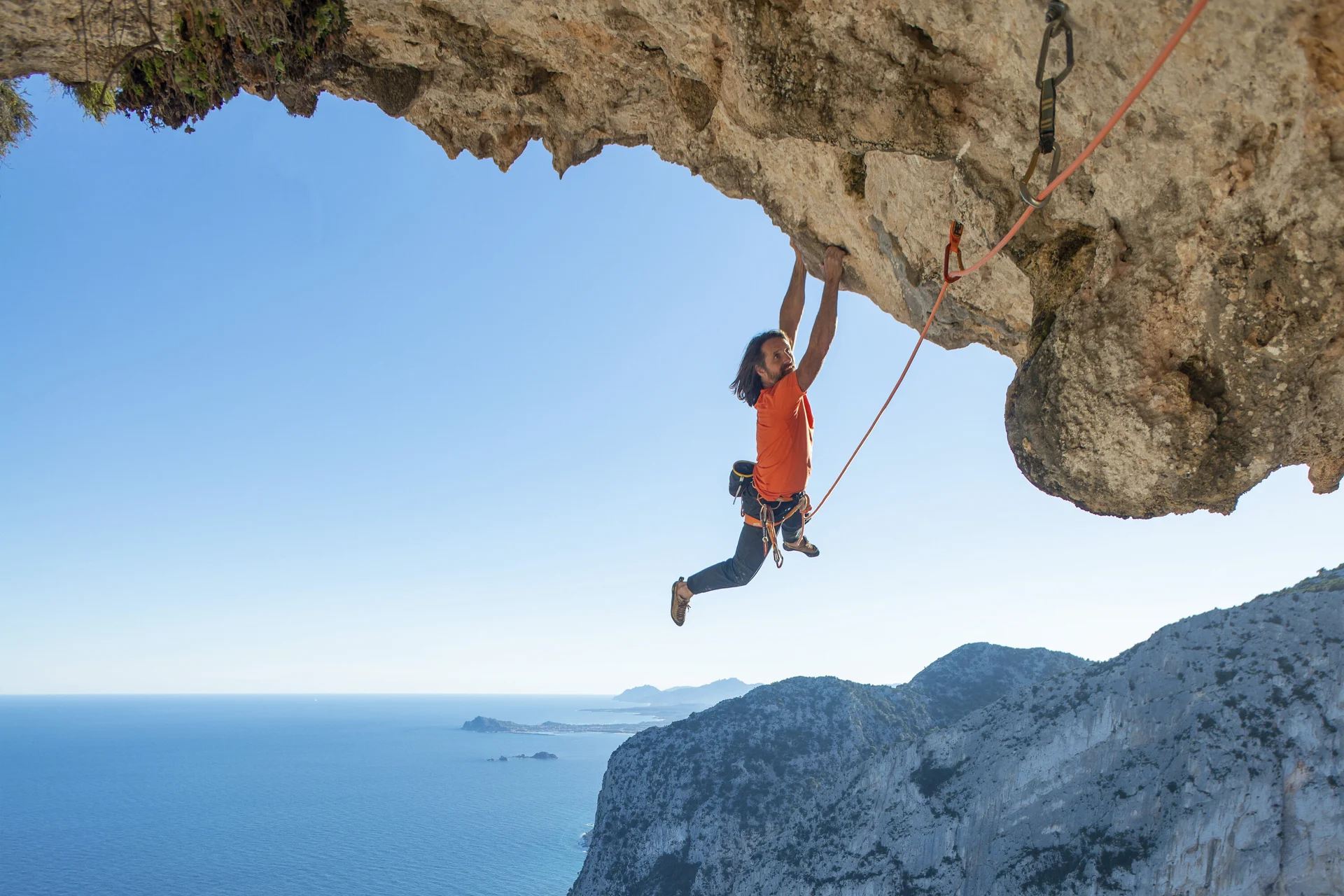 Mann hängt nur an den Armen im Überhang, im Hintergrund sieht man das Meer | © Alexander Huber