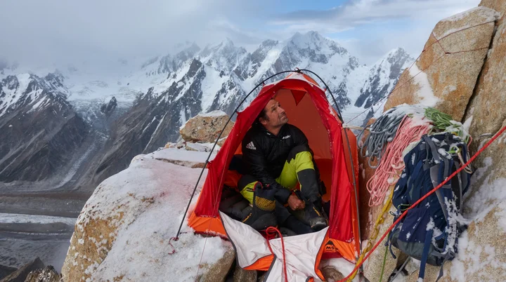 Mann hat kleines Zelt in der verschneiten Felswand aufgebaut, sitzt darin und blick nach oben. Um ihn herum liegen Seile. | © Alexander Huber