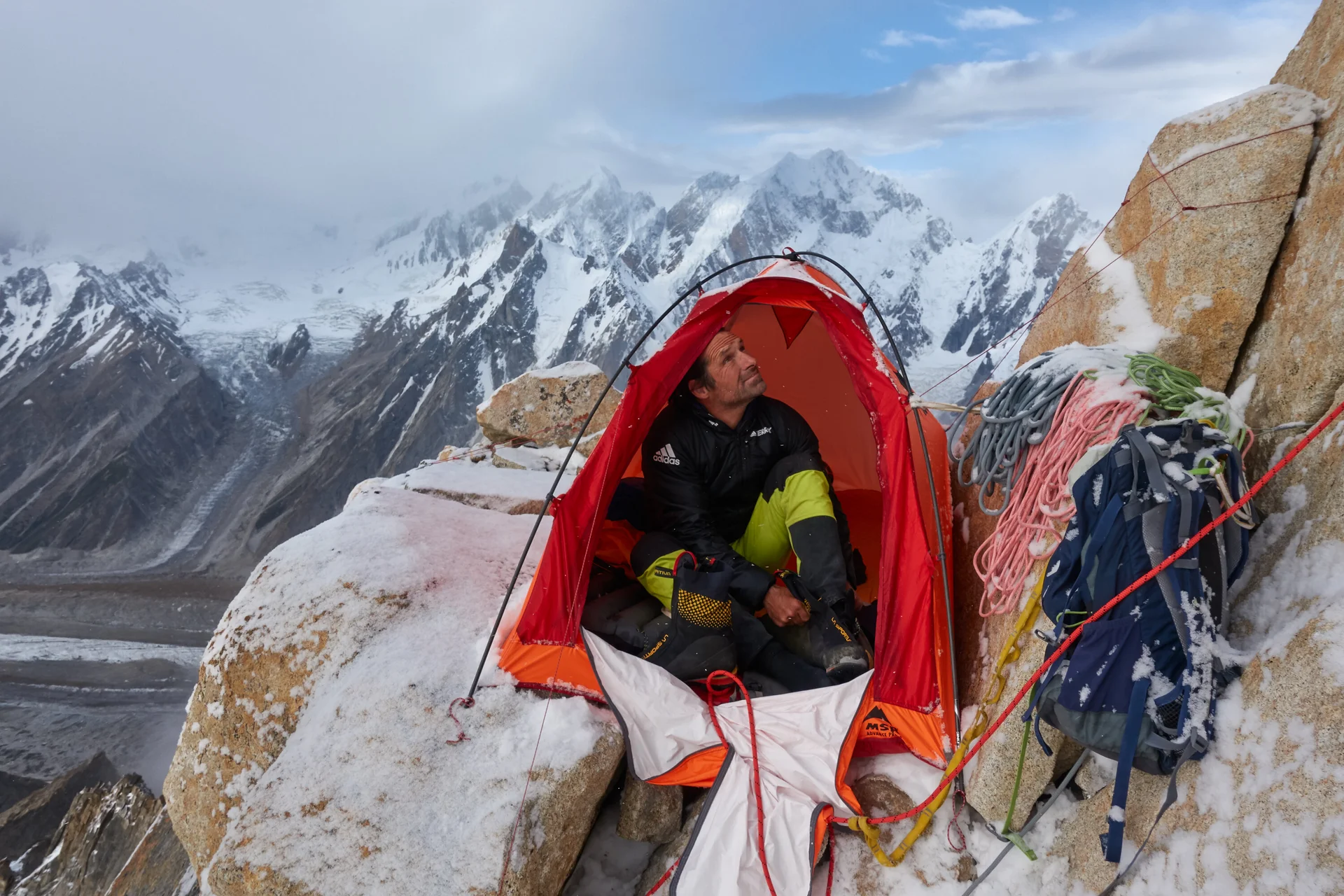 Mann hat kleines Zelt in der verschneiten Felswand aufgebaut, sitzt darin und blick nach oben. Um ihn herum liegen Seile. | © Alexander Huber