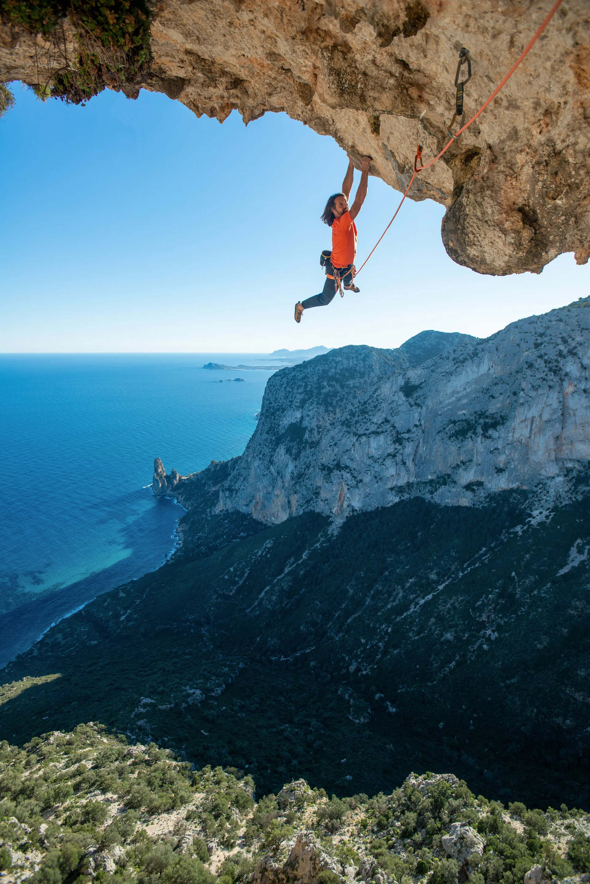 Kletterer hängt im Überhang, im Hintergrund sieht man das Meer | © Alexander Huber