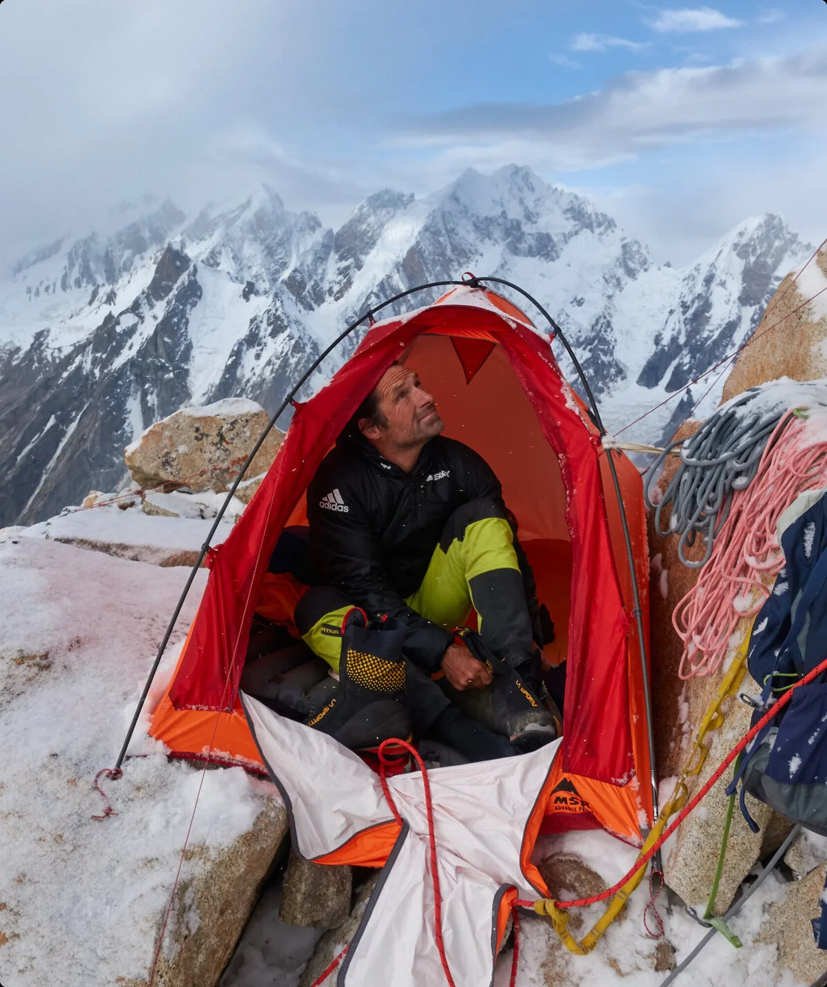 Mann hat kleines Zelt in der verschneiten Felswand aufgebaut, sitzt darin und blick nach oben. Um ihn herum liegen Seile. | © Alexander Huber