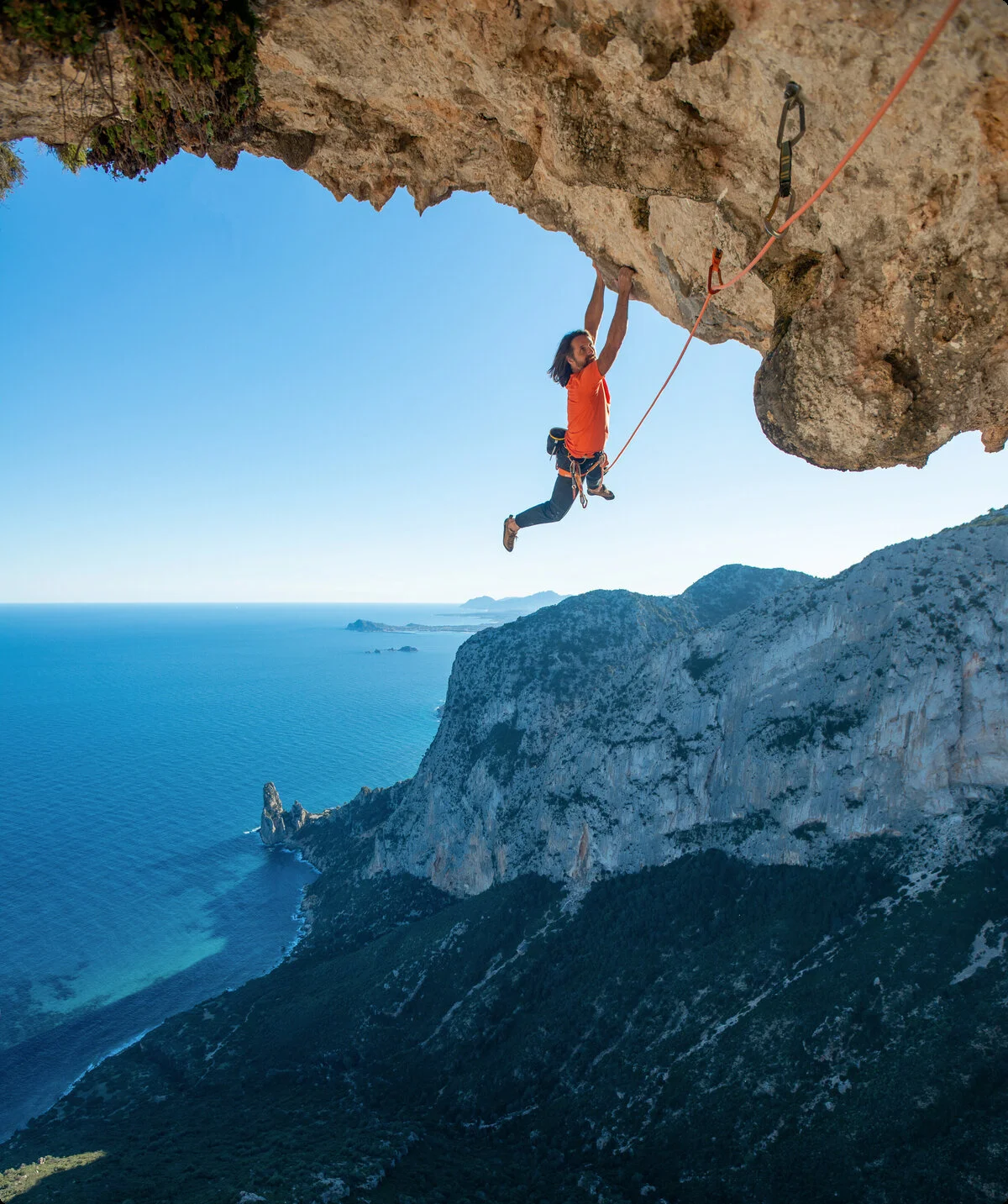 Kletterer hängt im Überhang, im Hintergrund sieht man das Meer | © Alexander Huber