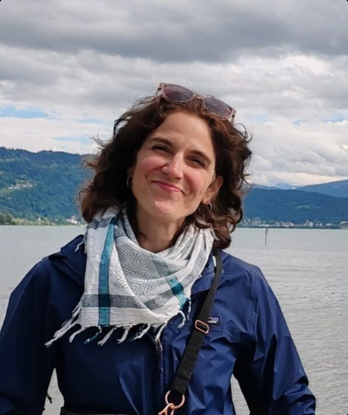 Portrait einer Frau mit dunklen Haaren, die ihre Sonnenbrille ins Haar gesteckt hat, im Hintergrund sieht man Wasser und Berge | © Stefanie Pausch