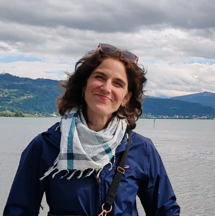 Portrait einer Frau mit dunklen Haaren, die ihre Sonnenbrille ins Haar gesteckt hat, im Hintergrund sieht man Wasser und Berge | © Stefanie Pausch