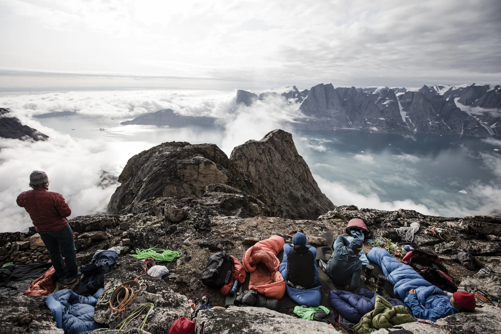 Schlafsäcke auf Felsvorsprung, im Hintergrund Wolken, ein Mann blickt in die Ferne | © © Stefan Glowacz GmbH