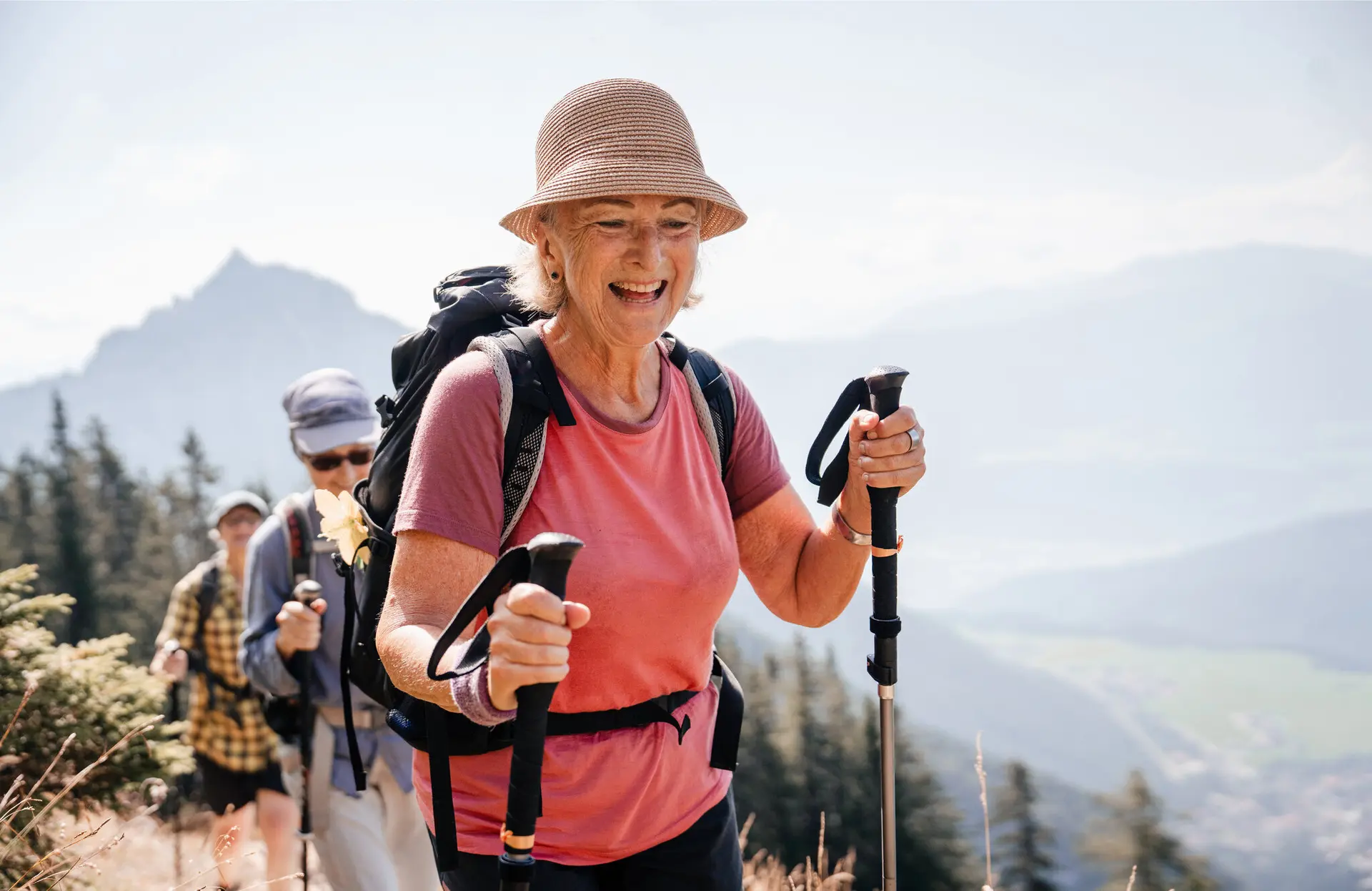 Eine ältere Frau mit Stöcken und Sonnenhut führt glücklich eine Wandergruppe an. | © DAV / Marisa Koch